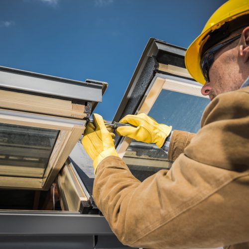 Caucasian Worker Wearing Safety Helmet and Protective Gloves Carrying Out Repair Works on Roof Skylight Windows Using His Screwdriver.